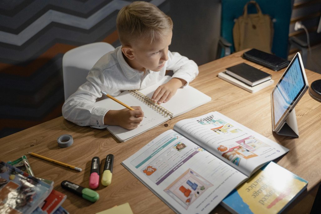 A focused young boy engages in distance learning with a tablet, books, and notebooks on a desk.